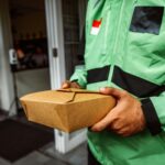 A deliveryman in a green jacket holding an eco-friendly food package outside a restaurant.